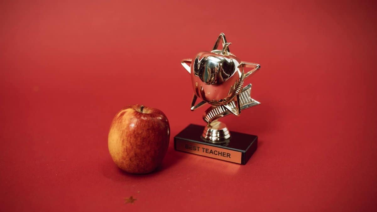 A 'Best Teacher' trophy alongside an apple on a vivid red background symbolizing education and recognition.
