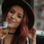 Smiling woman with red hair in a hat enjoying a glass of wine indoors.