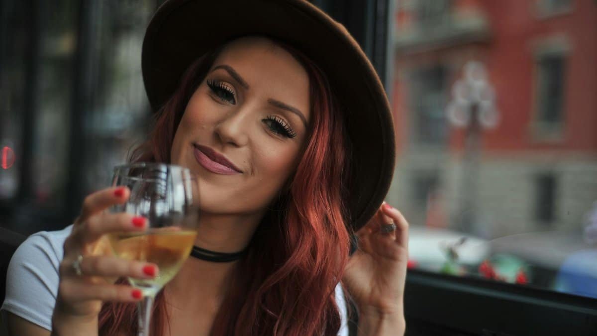 Smiling woman with red hair in a hat enjoying a glass of wine indoors.