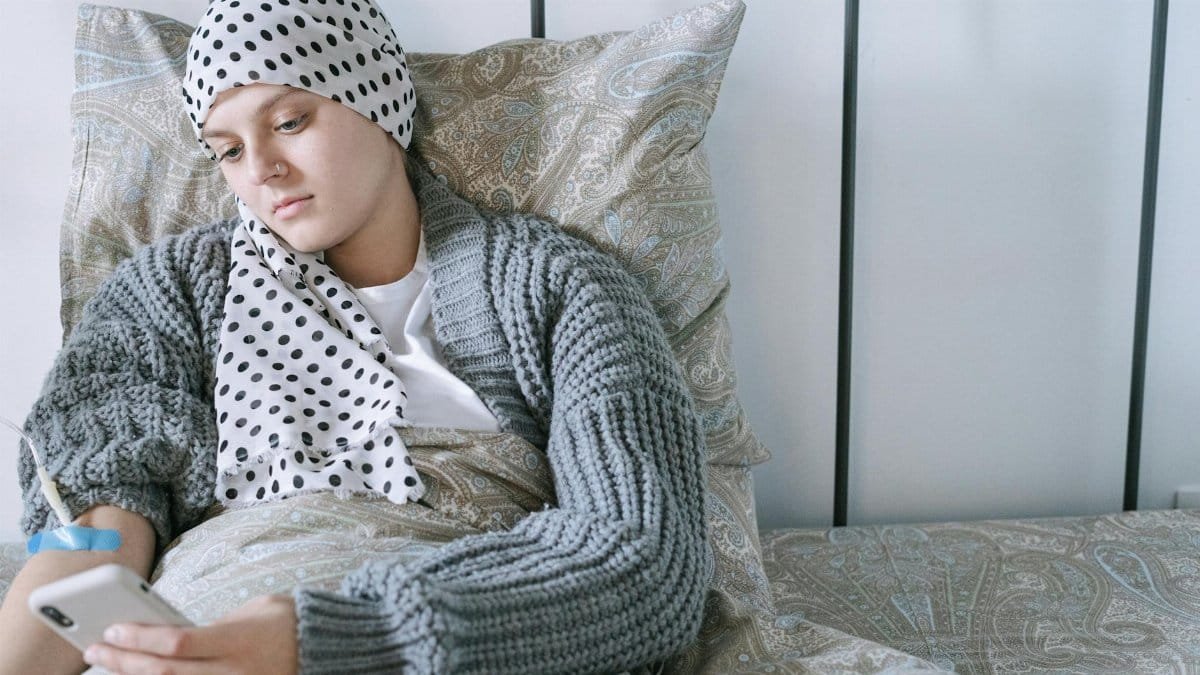 Woman resting in bed wearing a scarf, using her phone during cancer treatment.