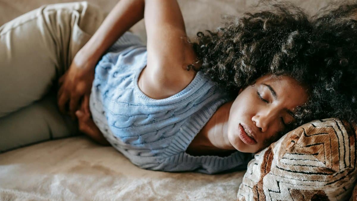High angle of African American woman with afro hairstyle and closed eyes having pain in belly lying on sofa