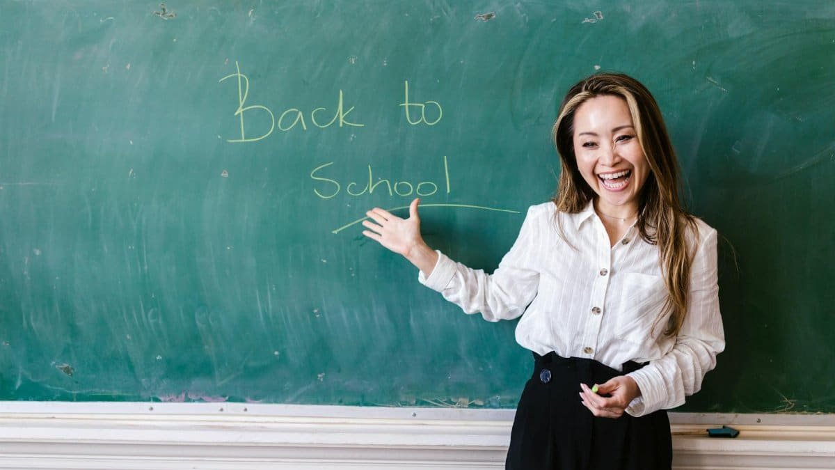 Smiling teacher in a classroom pointing at back to school message on a chalkboard.