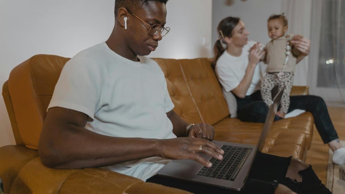 A man works on a laptop while a woman cares for a toddler on a sofa, highlighting multitasking at home.