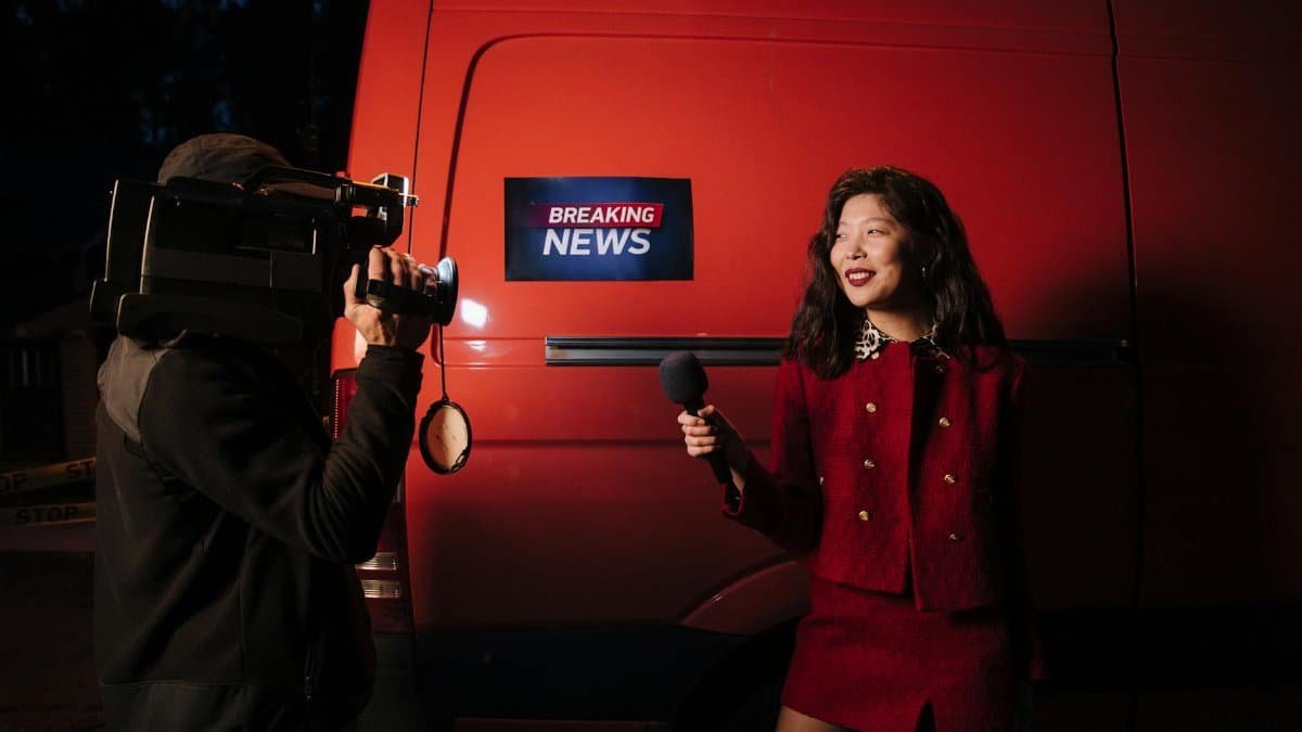 Dynamic news reporter holding microphone near red van during nighttime outdoor broadcast.