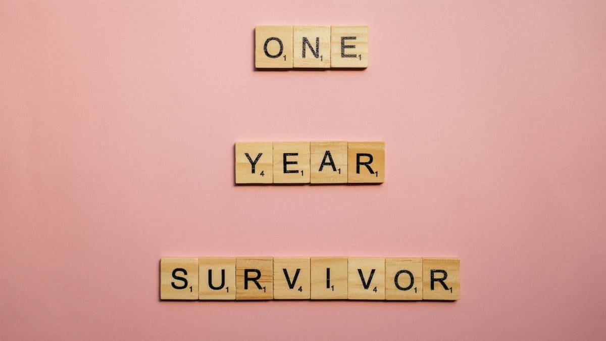 Wooden tiles spell 'One Year Survivor' on a pink background, symbolizing strength and resilience.