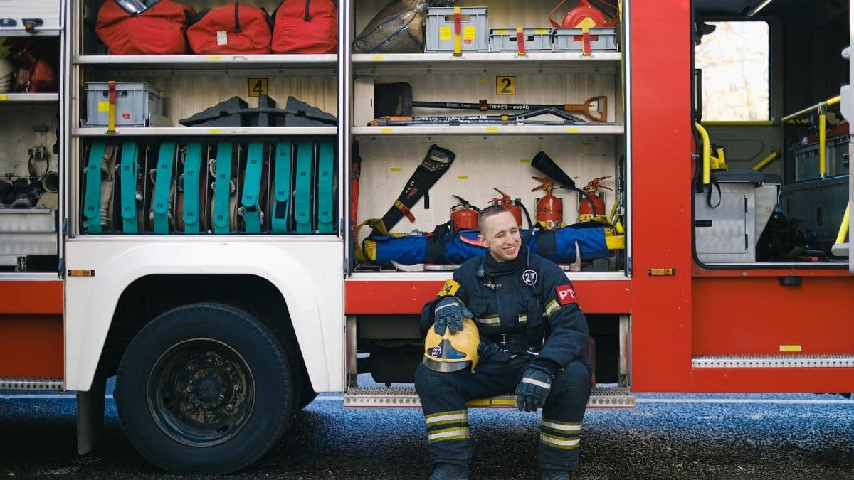 Firefighter in uniform sitting by fire truck, surrounded by equipment, smiling.