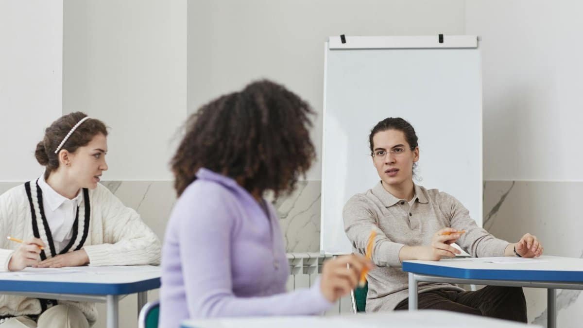 Three students engage in a group discussion in a well-lit, modern classroom setting.