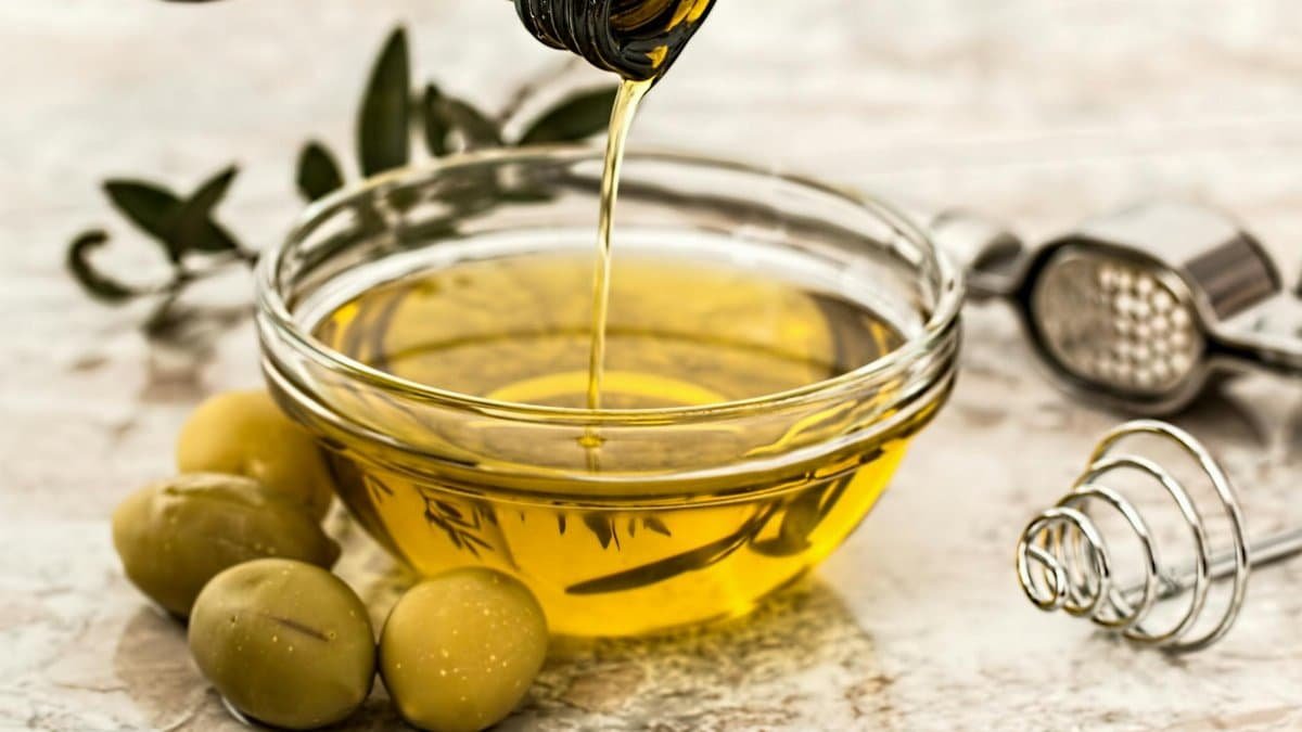 Close-up of olive oil being poured into a glass bowl surrounded by fresh olives and kitchen tools.