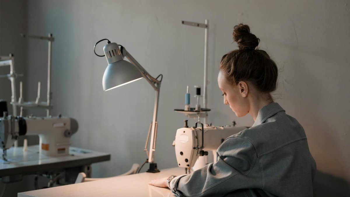 A woman seamstress focusing on her work with a sewing machine under a lamp in a tailor studio.