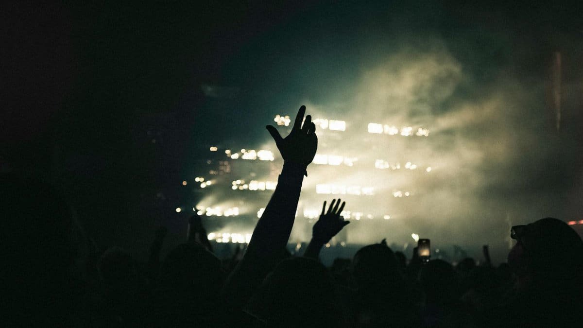 Silhouettes in concert crowd with raised hands against dramatic stage lights.