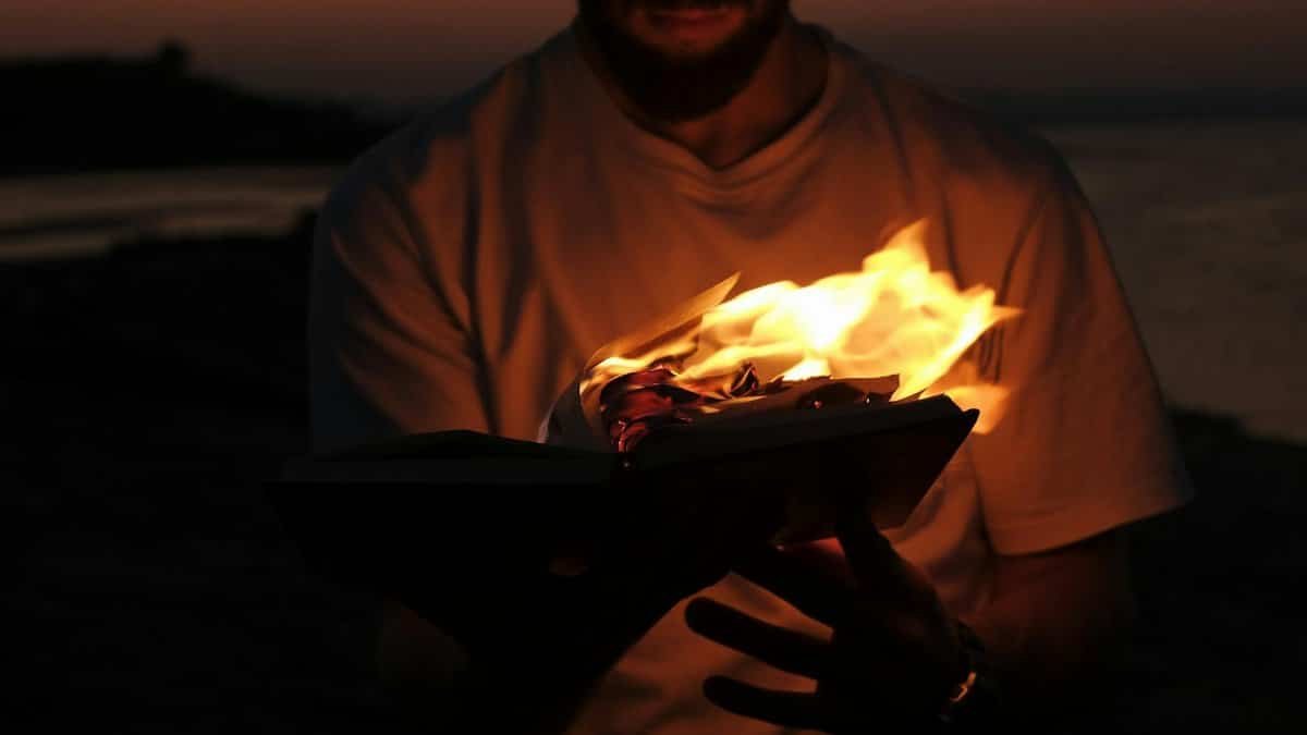 A man in a white shirt holds a flaming book by the water at sunset, creating a dramatic scene.