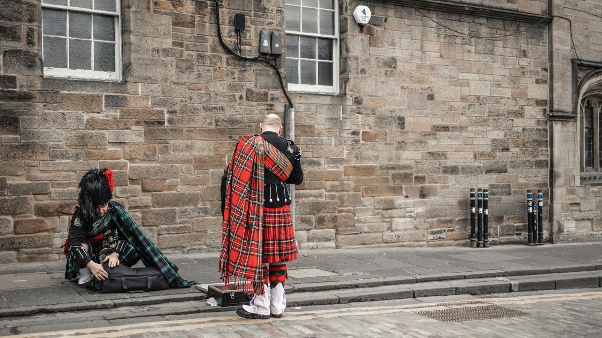 Two bagpipers in traditional Scottish kilts prepare instruments on a cobblestone street.