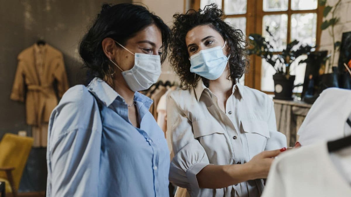 Two women wearing masks shopping in a clothing boutique, choosing outfits together.