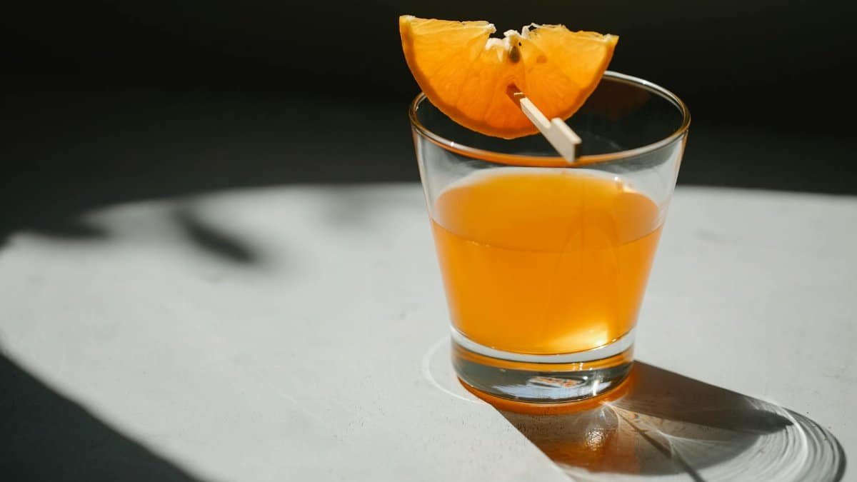 Glass of alcohol drink decorated with slice of orange on wooden stick placed on white table on blurred black background