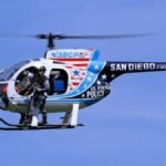 San Diego police helicopter with armed officer in flight against clear blue sky.