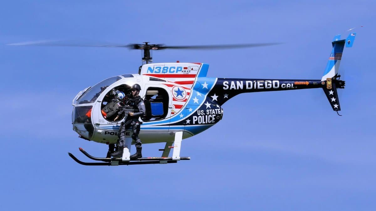 San Diego police helicopter with armed officer in flight against clear blue sky.