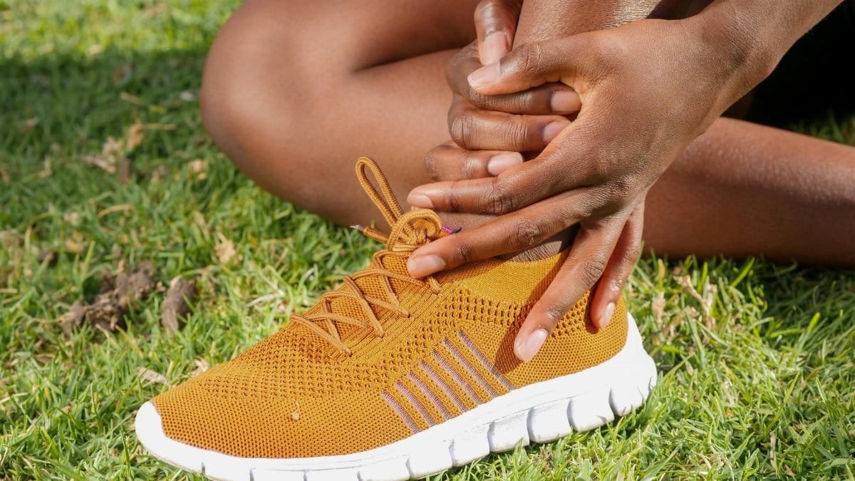 Close-up of athlete holding ankle with running shoe on grass, indicating pain or injury.