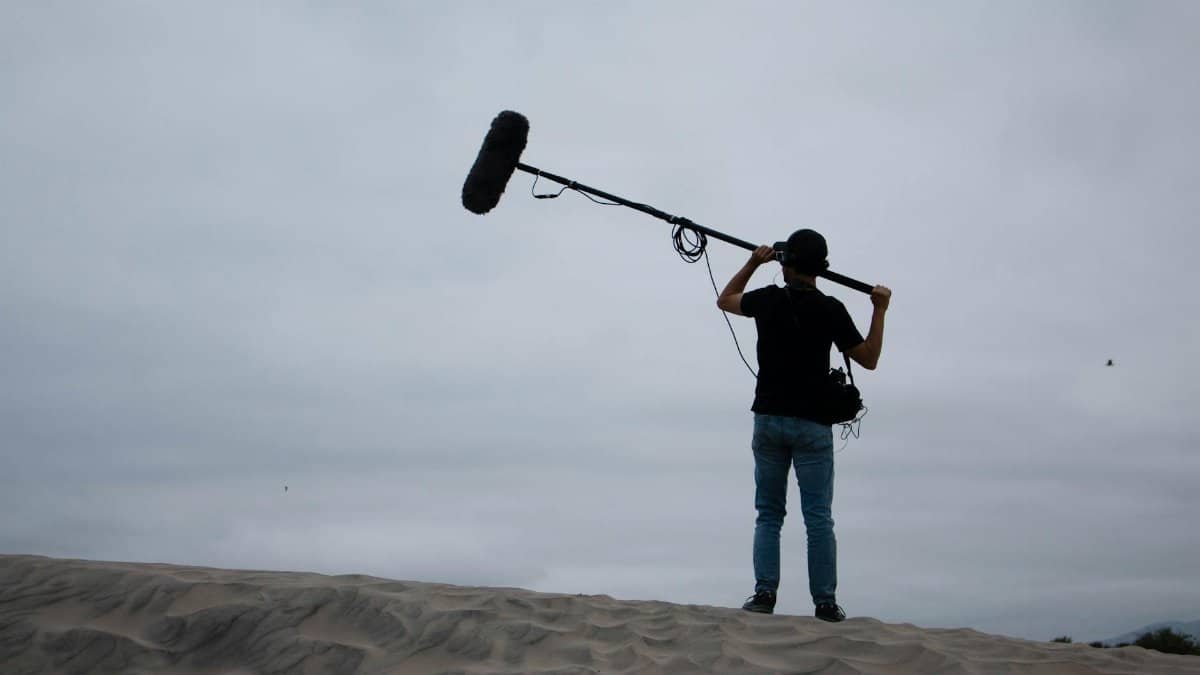 Back view of sound technician with boom mic on sandy dunes, working outdoors.