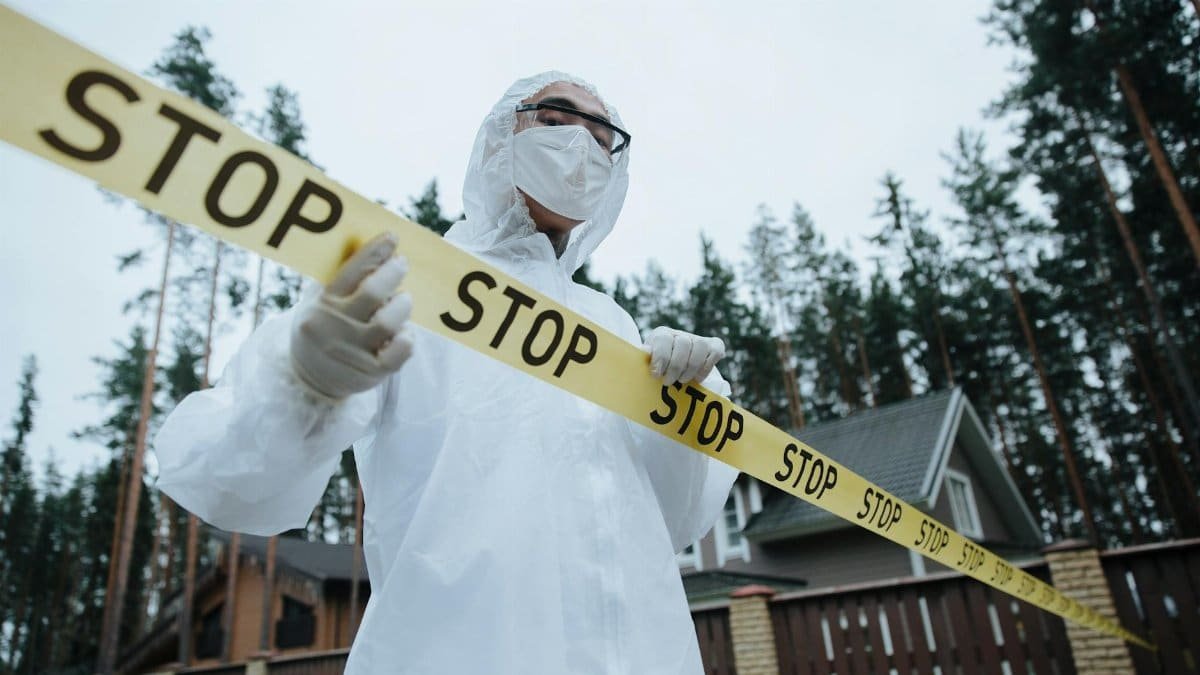 A forensic investigator in protective gear handles caution tape by a house in a wooded area.