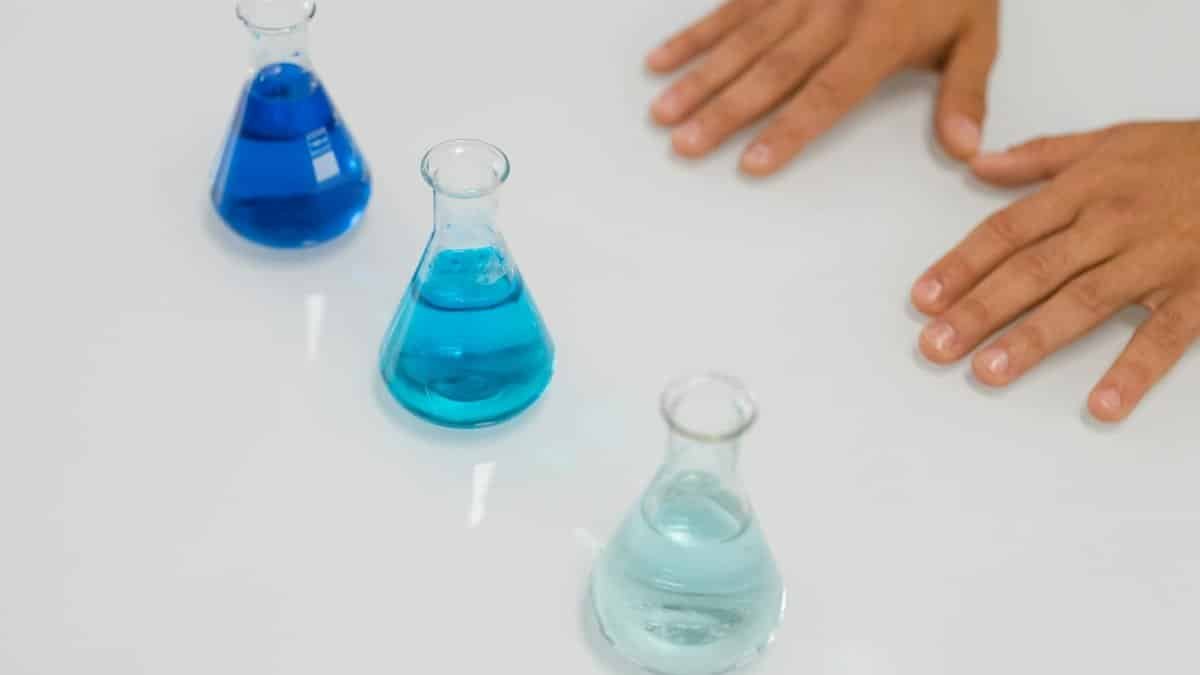 Blue liquids in Erlenmeyer flasks on a lab table with hands visible, symbolizing science research.
