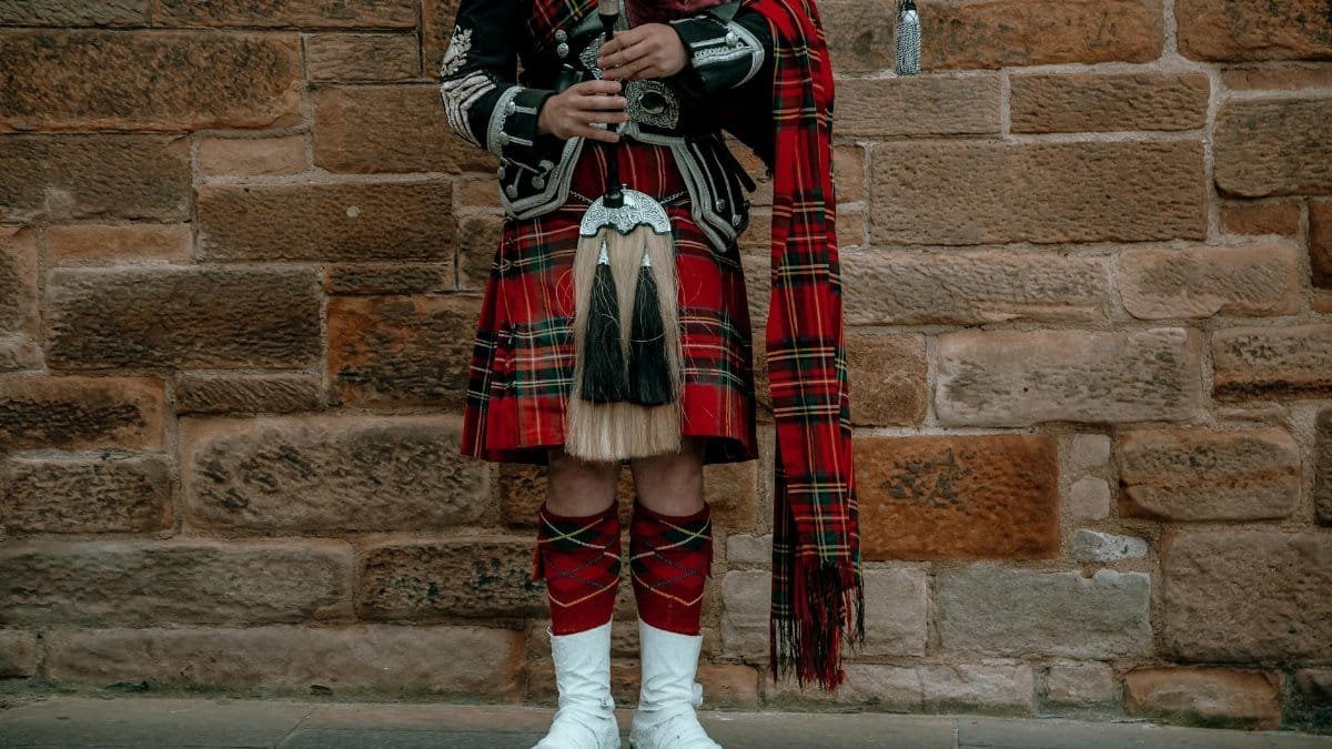 A Scottish bagpipe player in traditional attire with kilt against a stone wall.