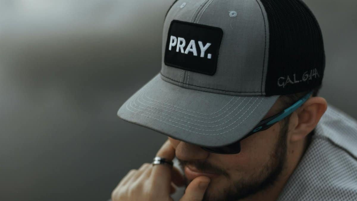 A man with sunglasses and beard wears a cap labeled 'Pray', symbolizing calm and meditation.