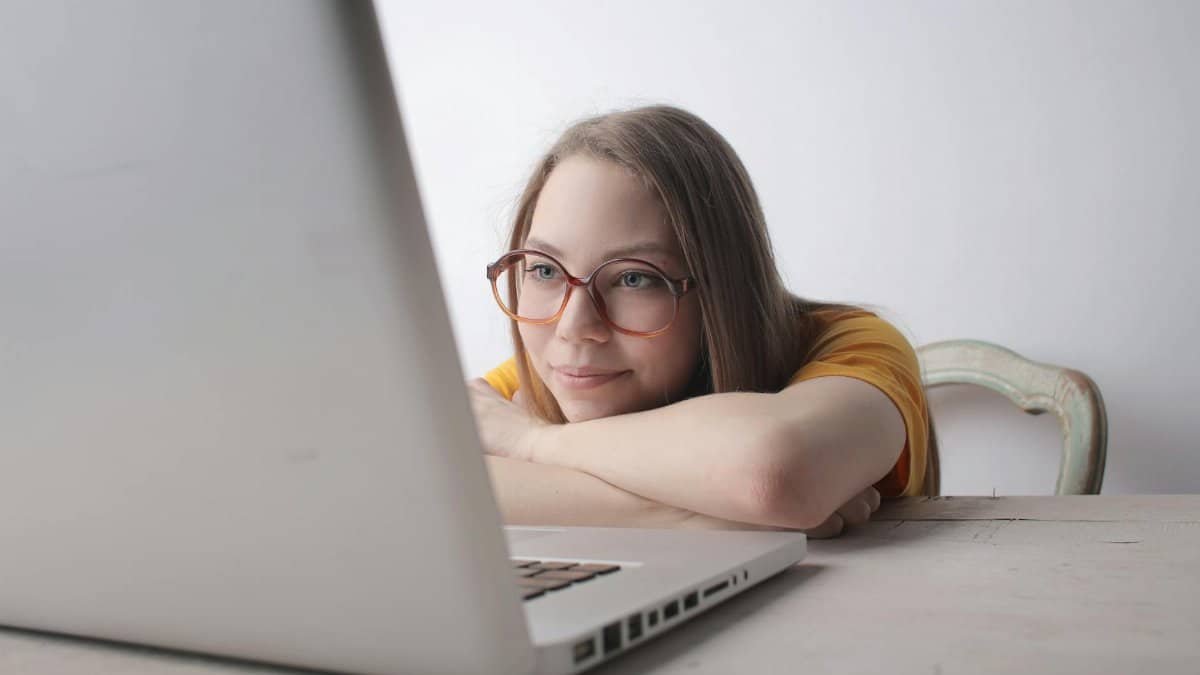 A young woman wearing eyeglasses relaxes at home, leaning on a table while using a laptop.