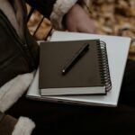 A person sitting outdoors with a notebook and laptop, surrounded by autumn leaves, representing a cozy work setup.