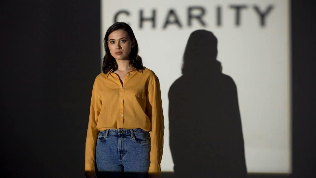 Asian woman in orange shirt, shadow cast against charity sign, indoors.
