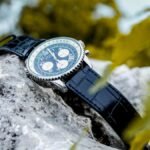 Elegant wristwatch with blue strap placed on a rock surrounded by vibrant foliage.