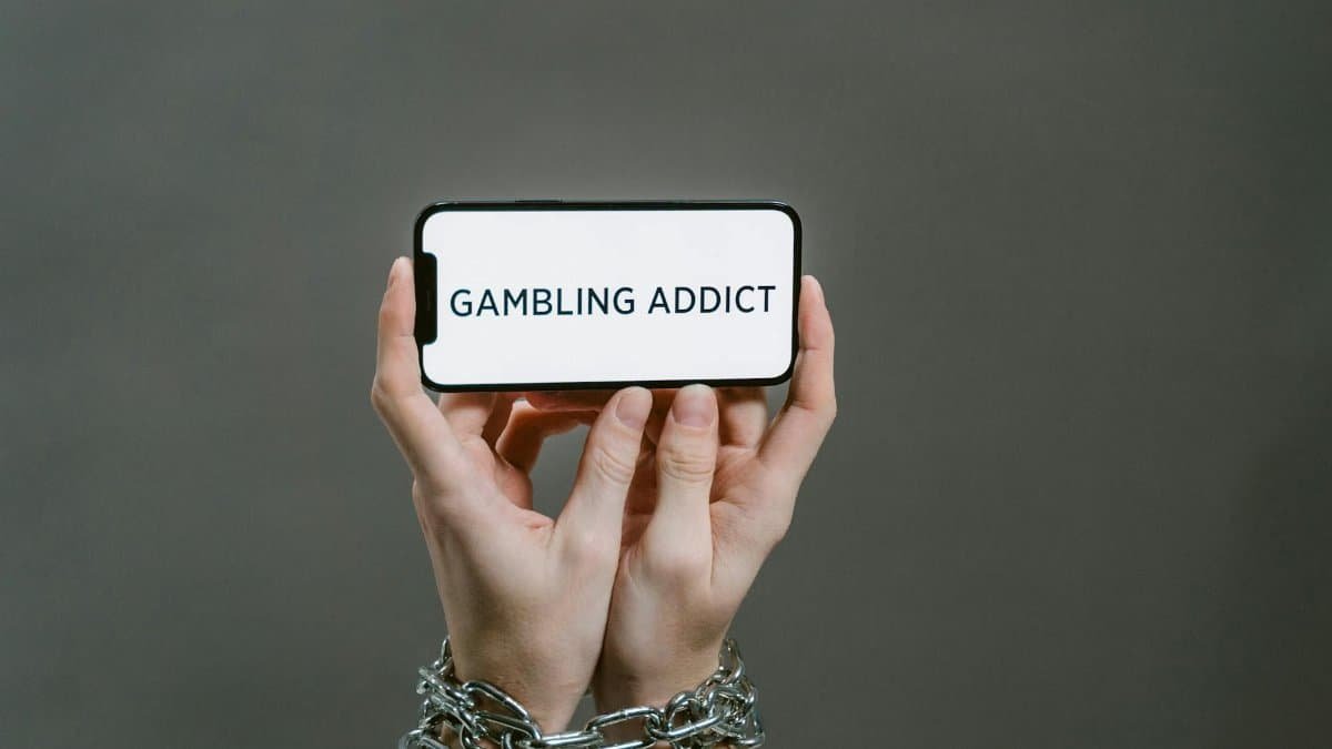 Chained hands hold a phone displaying the text 'Gambling Addict', highlighting addiction.