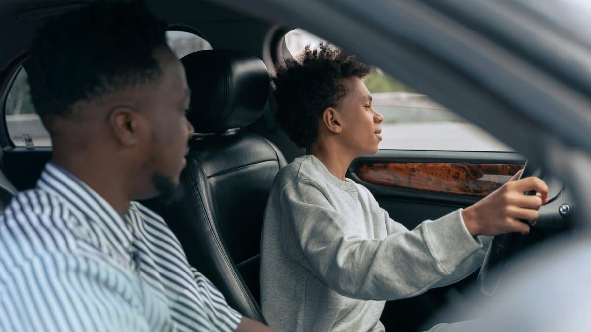 A father teaches his son to drive, sitting together in a car and discussing techniques. Side view indoors.