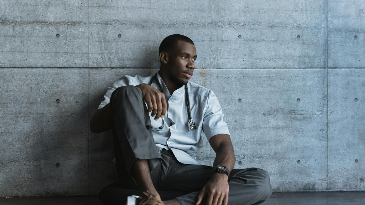 A thoughtful doctor in scrubs sits on a concrete floor, suggesting a moment of rest and reflection.