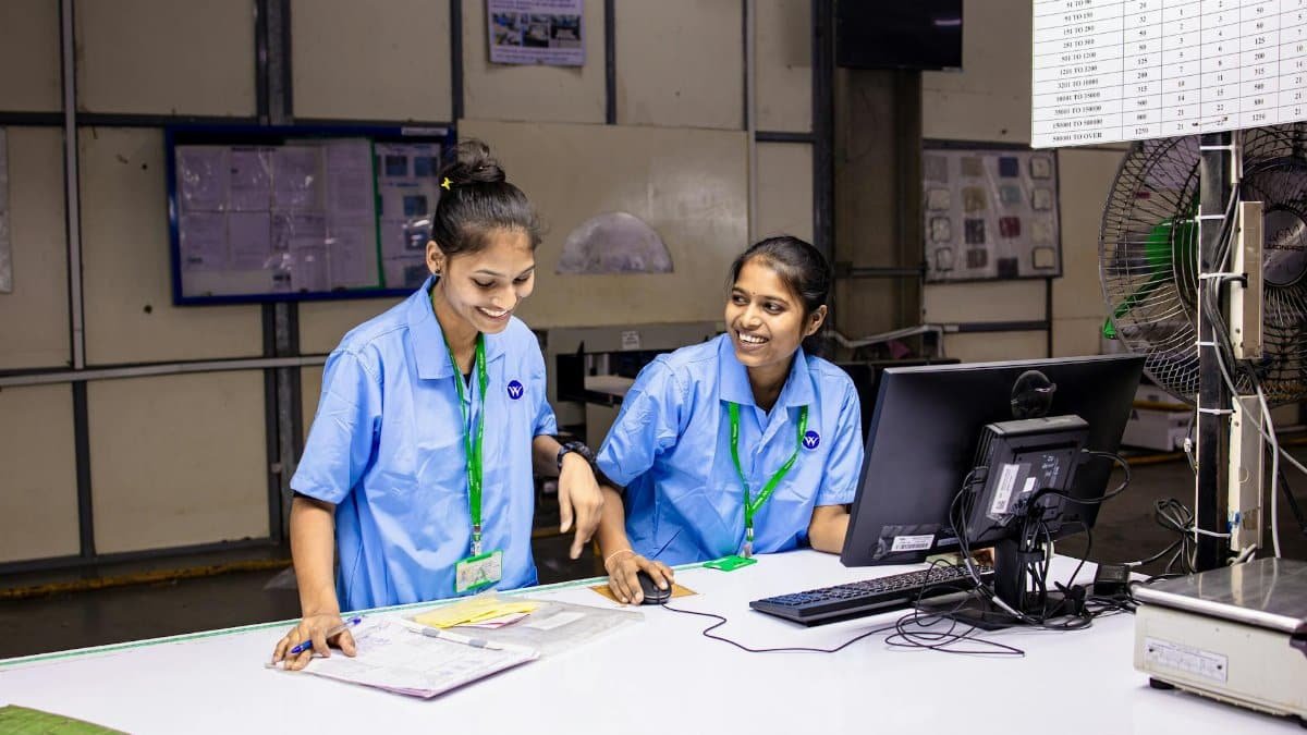 Two female workers discussing tasks at a factory workbench, smiling and engaged.