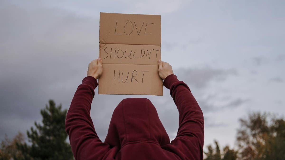 An anonymous person holds a cardboard sign reading 'Love Shouldn't Hurt' outdoors, advocating against violence.