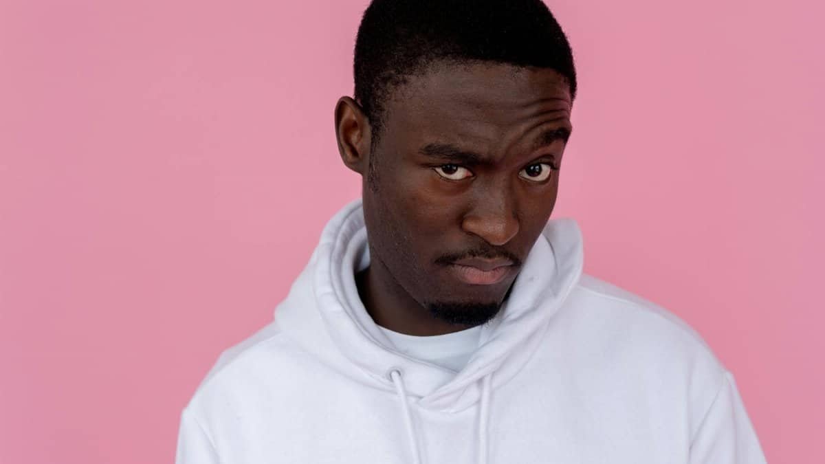 Serious African American male model wearing white sweatshirt looking at camera with unsure gaze against pink background