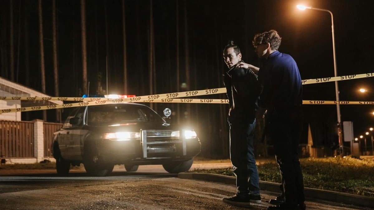 Police officer detaining a suspect near a crime scene at night with tape and patrol car.