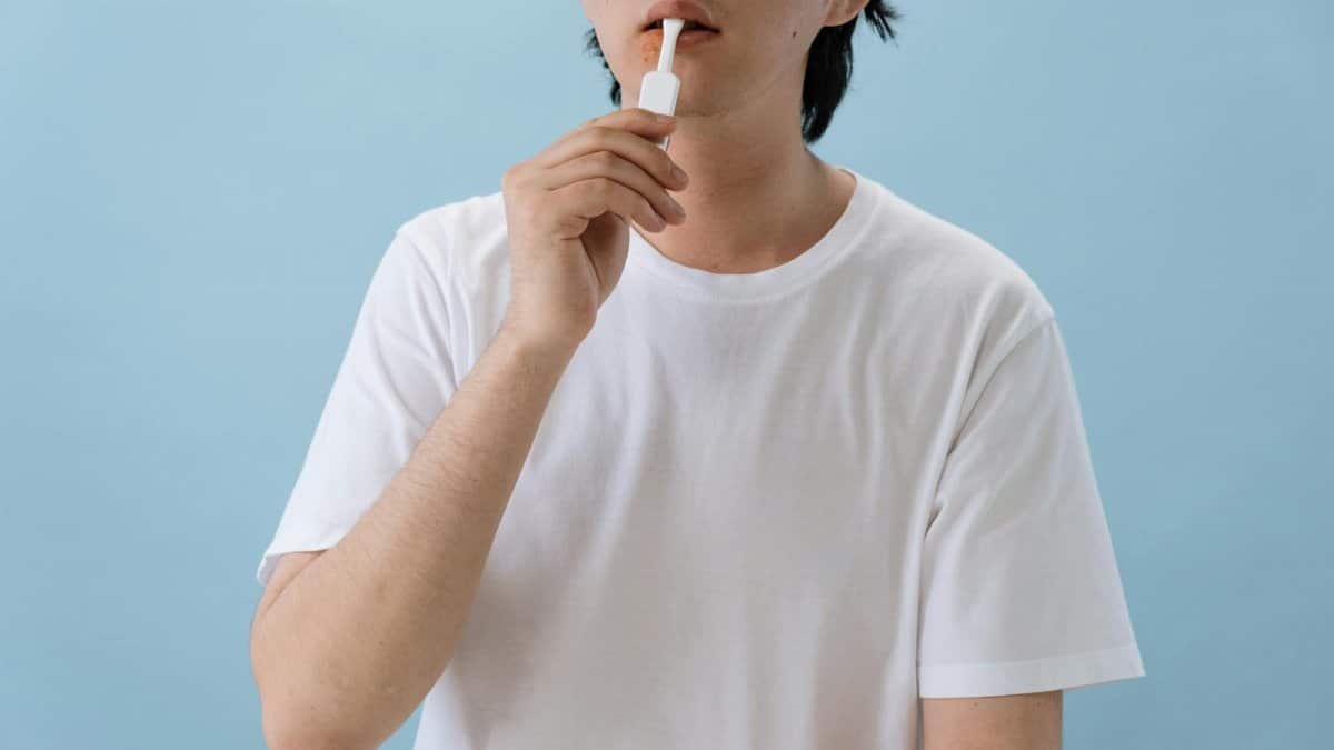 A person in white T-shirt using an oral swab test kit indoors with a blue background.