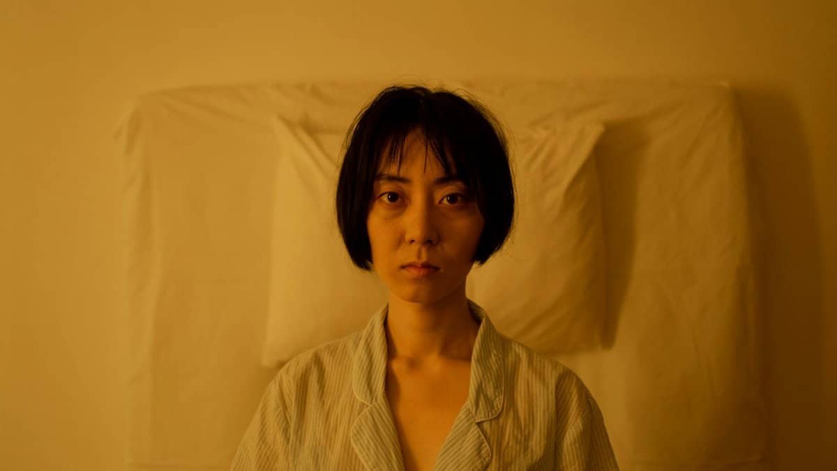 An Asian woman sits in bed with an expression of insomnia and stress, enveloped in a yellow ambiance.