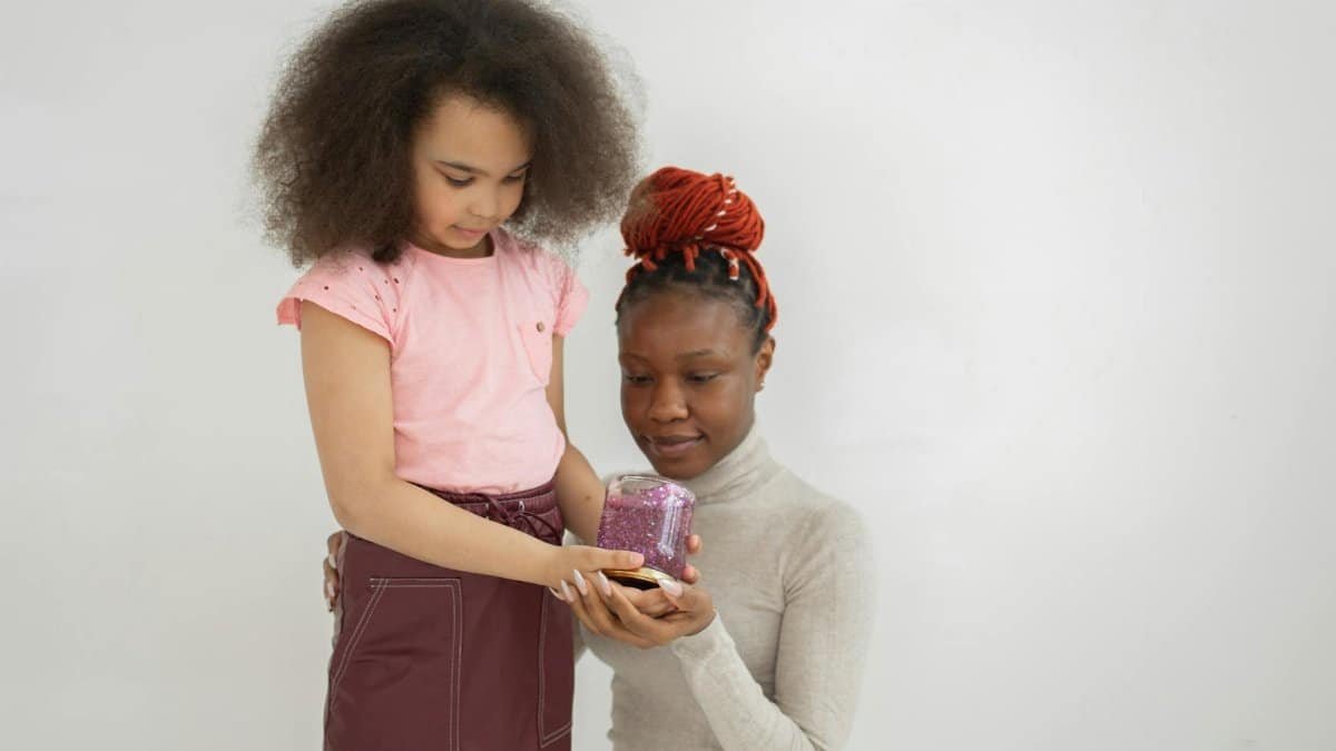 Cute African American little girl with dark curly hair standing near white wall and showing mother jar with handmade slime