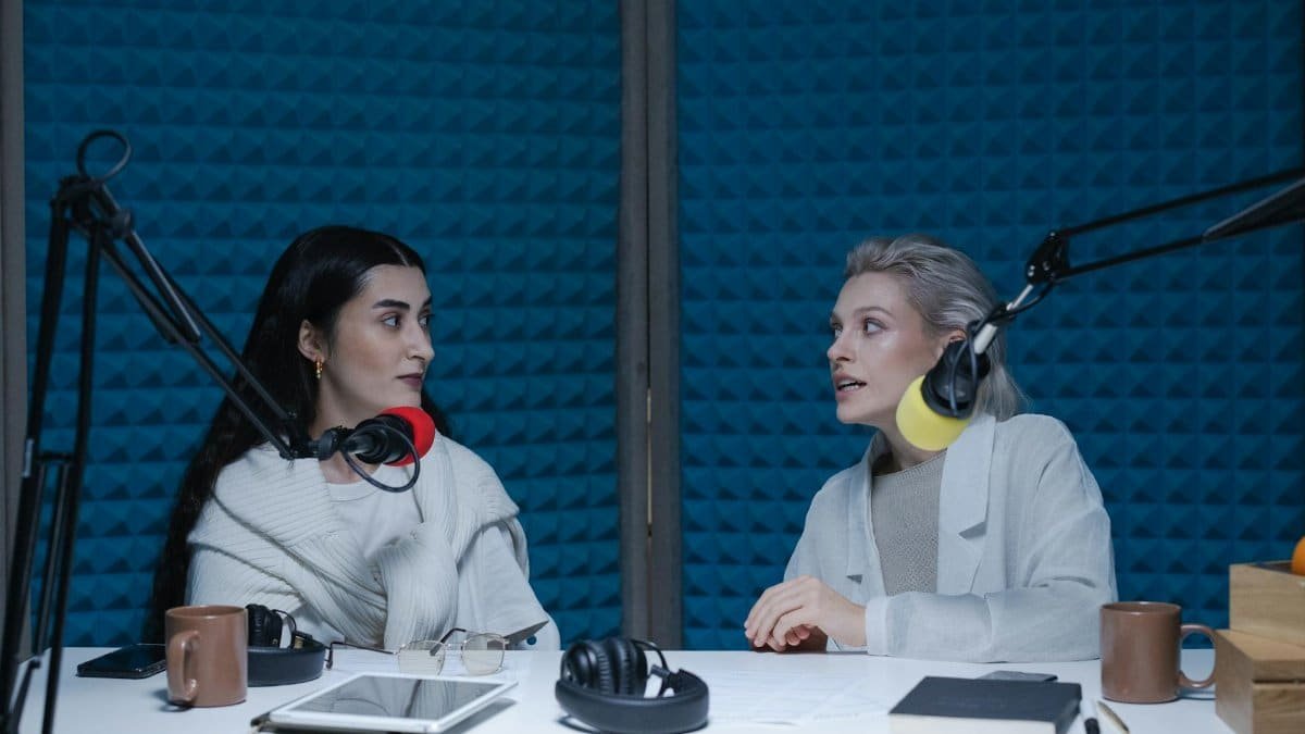 Two women engaged in conversation, recording podcast in studio with microphones and headphones.