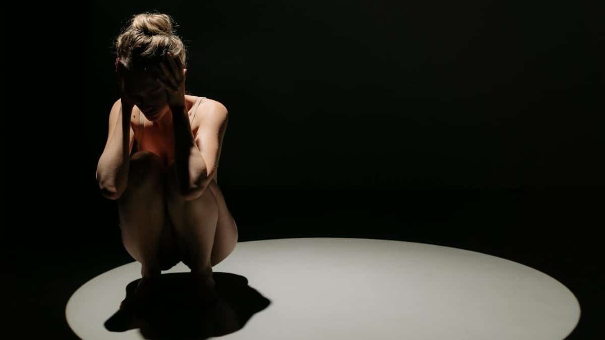 A woman crouching under a spotlight with hands on head, depicting stress.