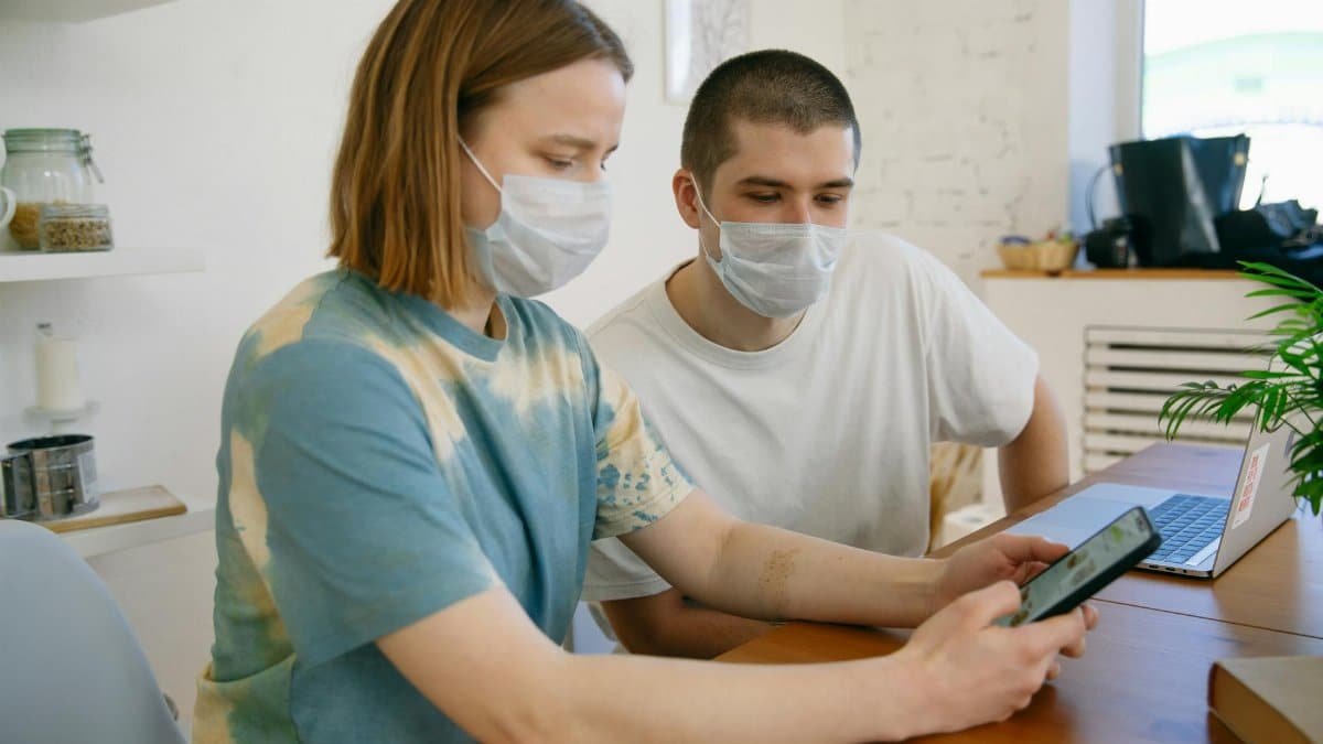 A couple with face masks using a smartphone indoors, embodying the new normal.