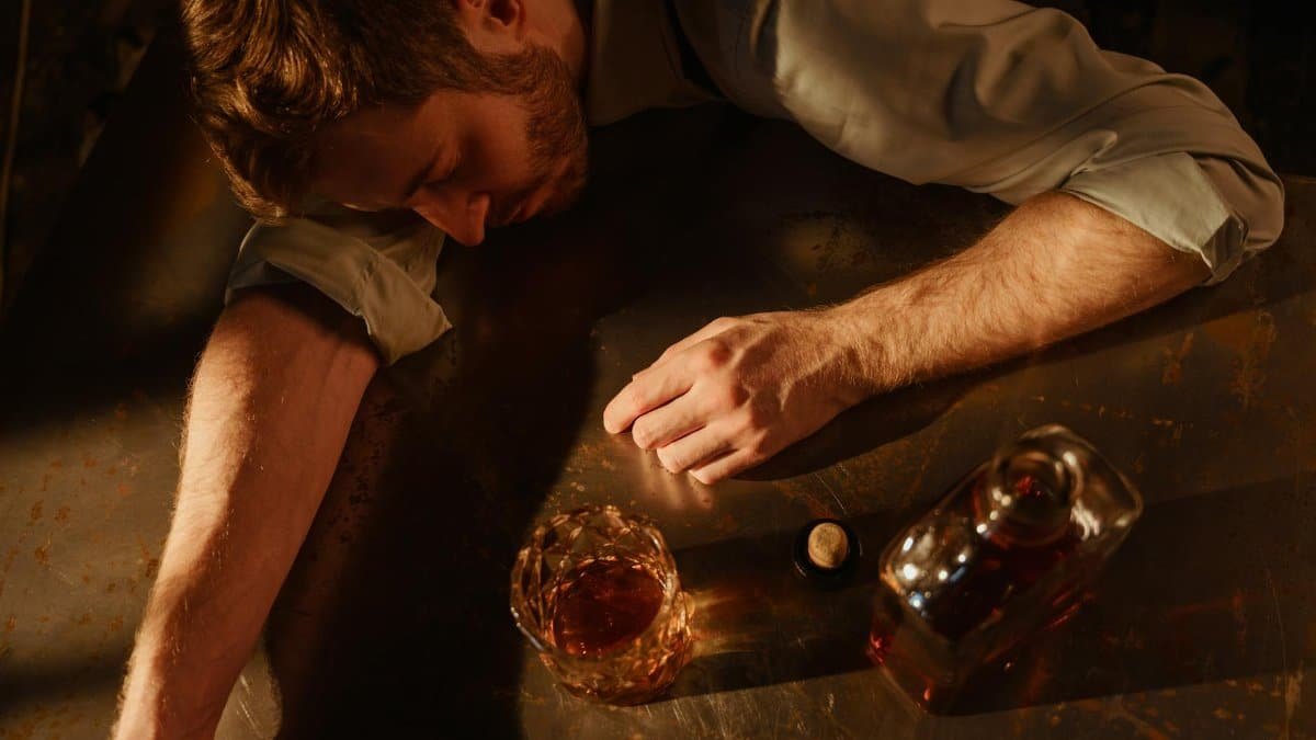 A man sleeps on a table with a whiskey glass and bottle nearby, suggesting exhaustion.