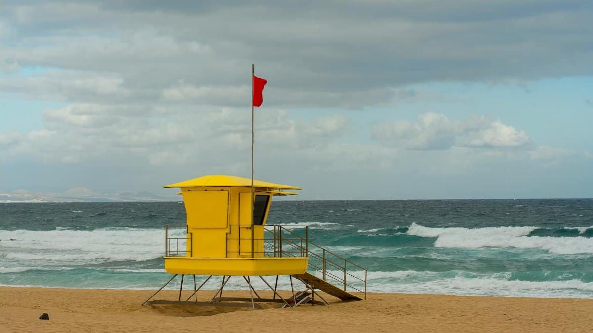 Yellow lifeguard tower with red flag on a sandy beach in Lanzarote, under cloudy skies.