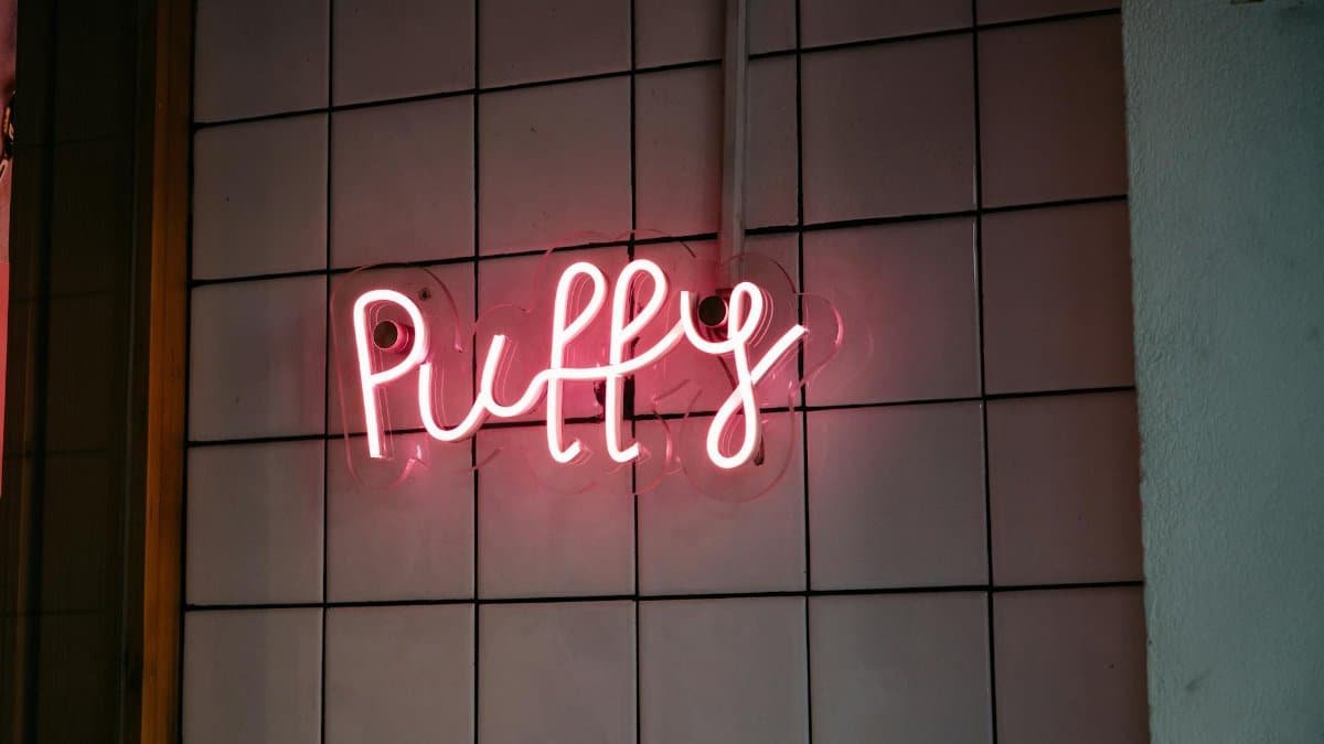 Pink neon 'Puffy' sign glowing against white tiled wall indoors.