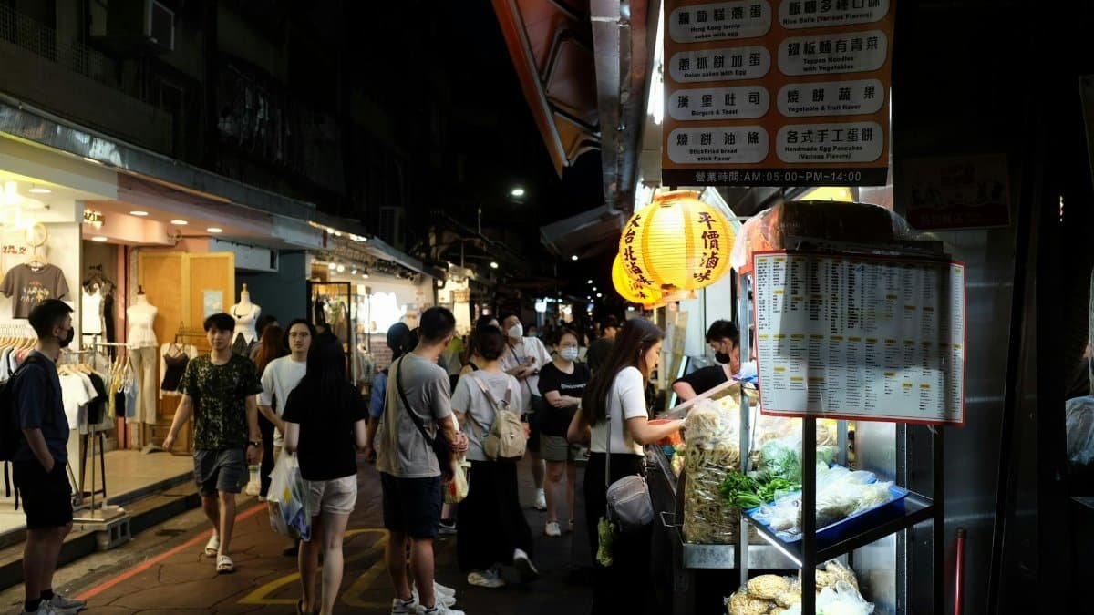 Crowded night market scene with people shopping and dining under bright lanterns.
