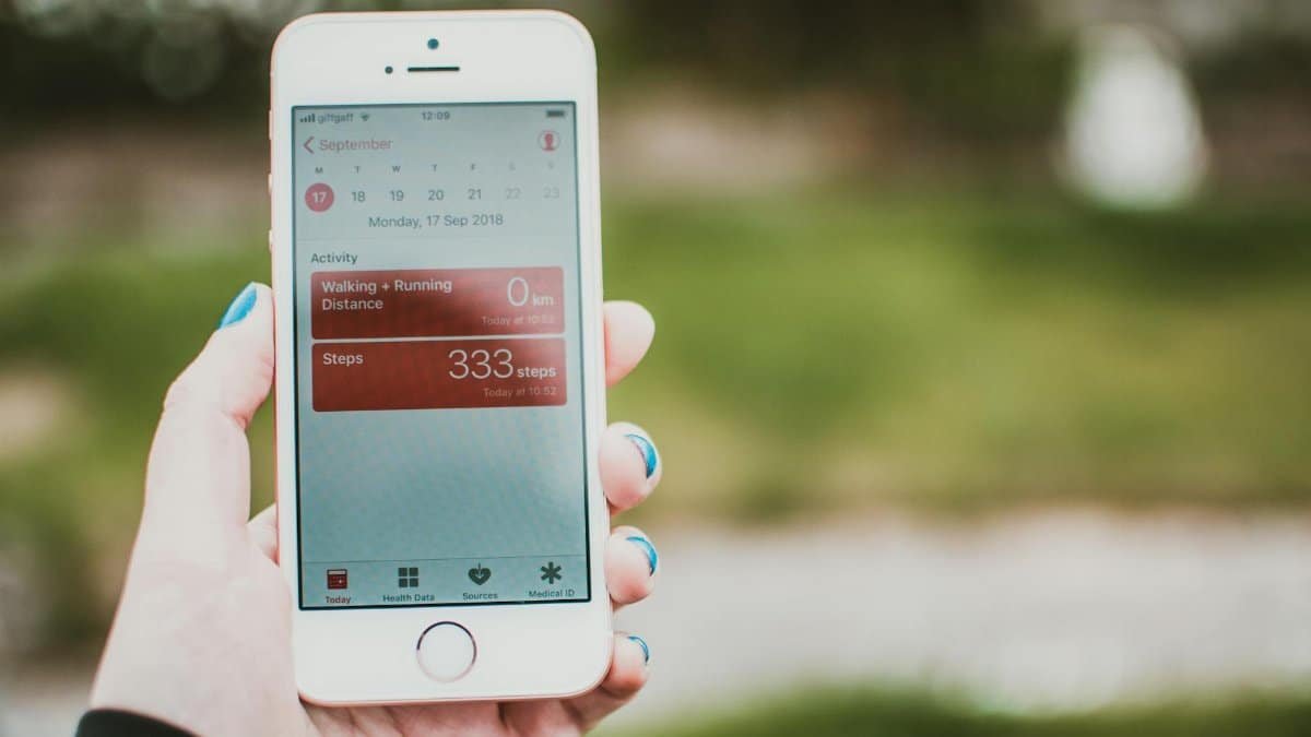 A person holds a smartphone showing a fitness app with step count and distance tracking outdoors.