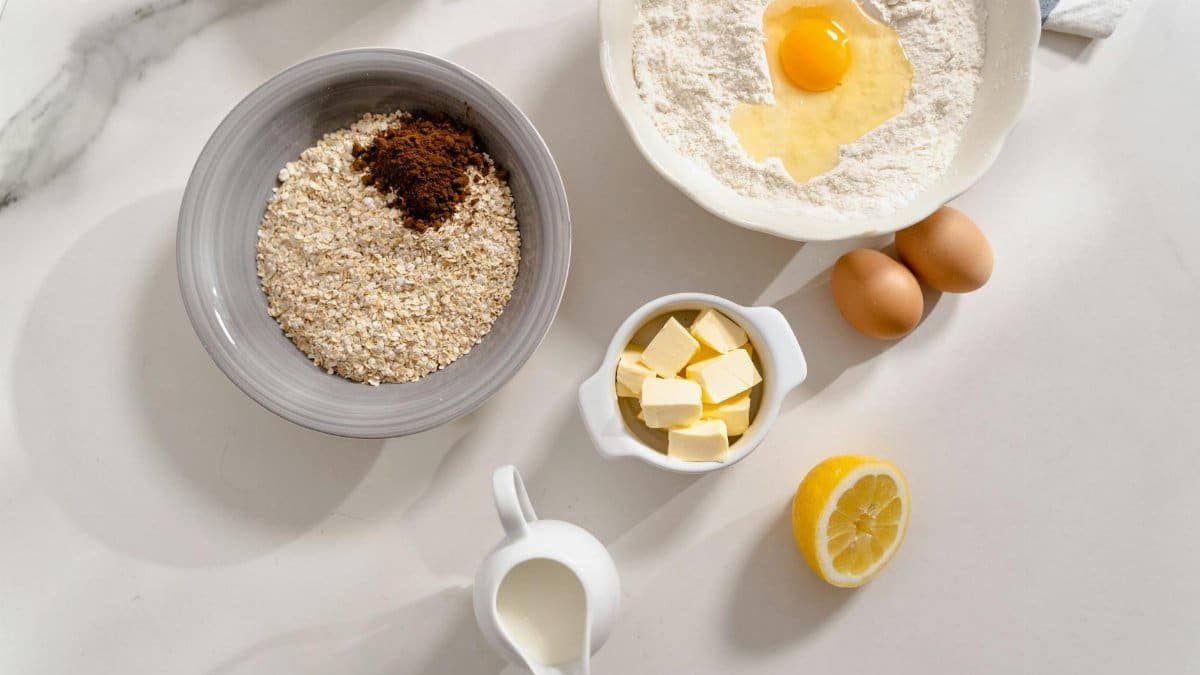 An overhead shot of baking ingredients including flour, eggs, butter, milk, and spices on a white counter.