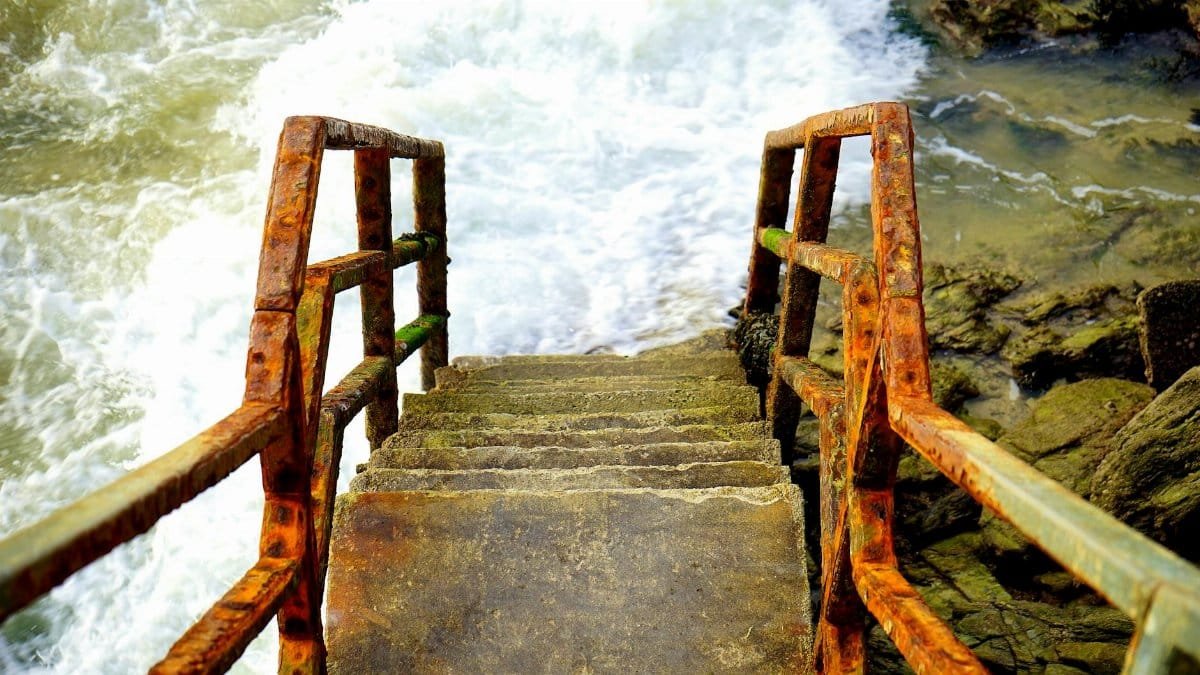 Rusted steps with railing descending to rough sea waves in Cornwall, UK.